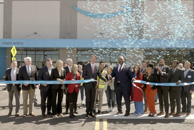 A ribbon-cutting ceremony at an Amazon facility featuring a group of people in business attire standing in front of the main entrance. Confetti is released as two individuals cut the blue ribbon with Amazon logos.