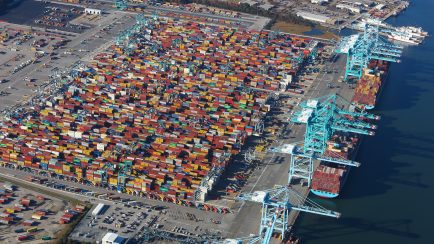 Aerial view of a busy shipping port with numerous colorful shipping containers stacked neatly in rows and large blue cranes along the waterline.